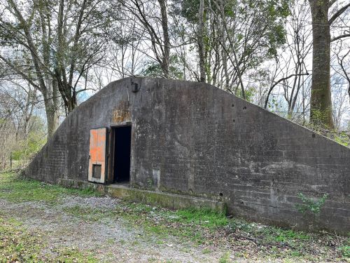 A weathered concrete structure with an orange door, surrounded by trees and gravel, set in a natural landscape.