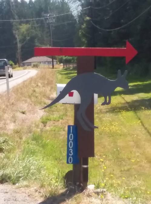 A wooden mailbox post with a kangaroo silhouette and a red arrow pointing right, located by a rural road.
