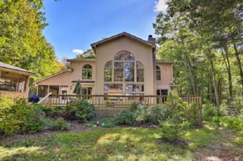 A large, modern house with a wooden deck, surrounded by trees and greenery under a clear blue sky.