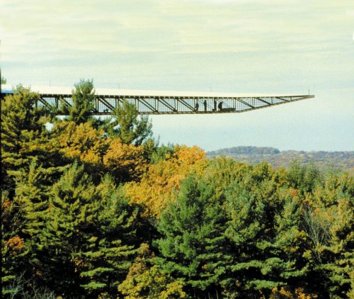 A long, narrow bridge extends over a forested landscape with colorful autumn trees under a cloudy sky.