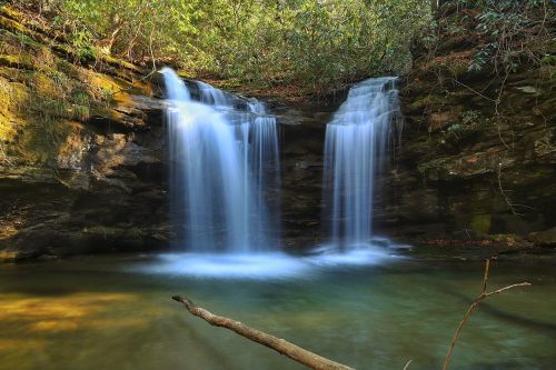 A serene waterfall cascades into a clear pool, surrounded by lush greenery and rocky cliffs.