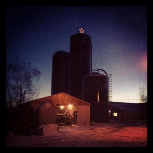 A silhouette of silos and a barn against a twilight sky, with a star atop one silo. Snow covers the ground.