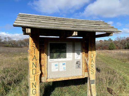 A wooden information kiosk in a grassy field, with "STANDING CEDARS" carved on the sides and a clear blue sky above.