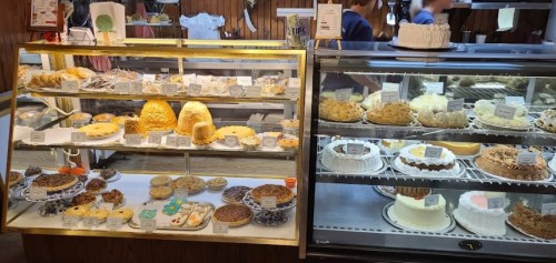 Display case filled with various cakes, pastries, and desserts, labeled with names and prices.