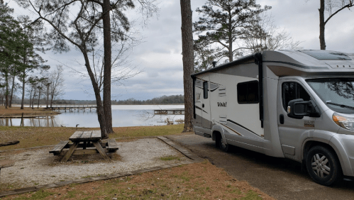 An RV parked by a serene lake, surrounded by trees and a picnic table nearby, under a cloudy sky.