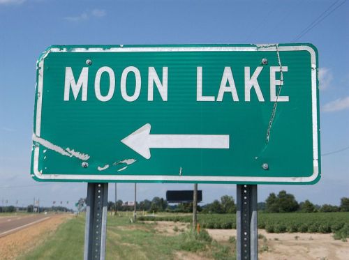 Green road sign directing left to Moon Lake, with a clear blue sky and fields in the background.