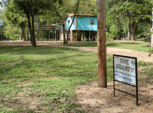 A sign for "The Shanty" near a blue cabin on stilts, surrounded by trees and grassy land.