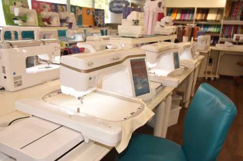 A row of sewing machines on tables in a brightly lit craft store, with colorful fabric rolls in the background.