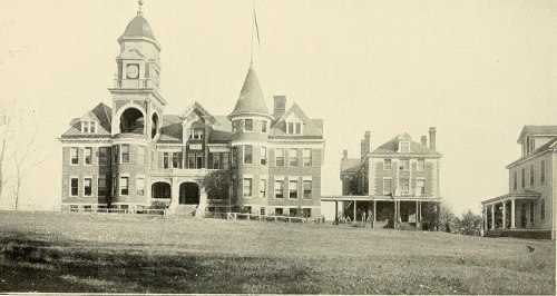 Historic building with a clock tower, turret, and large porch, set on a grassy hill. Black and white photo.