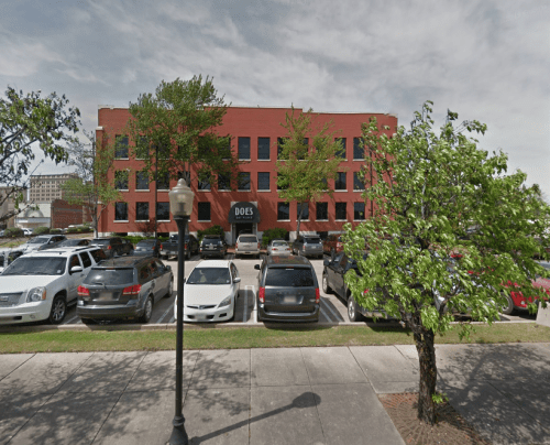 A red brick building with large windows, labeled "DOE'S," surrounded by parked cars and trees on a cloudy day.