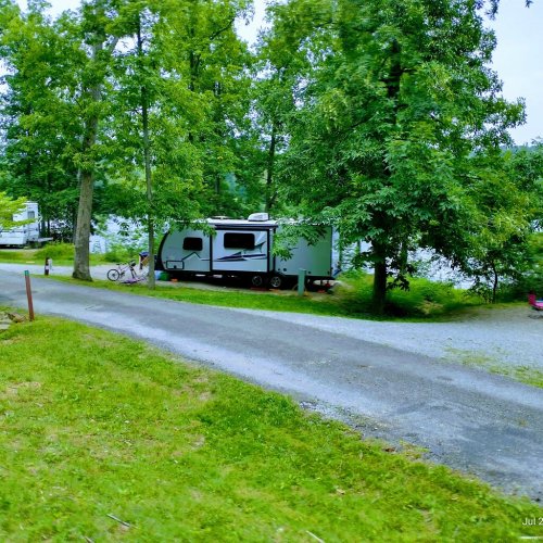 A gravel path leads to a parked RV surrounded by trees and a lake in the background.