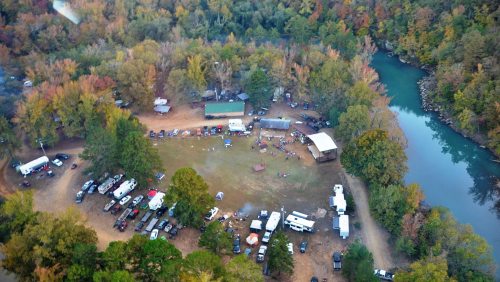 Aerial view of a campsite by a river, surrounded by colorful autumn trees and parked vehicles.