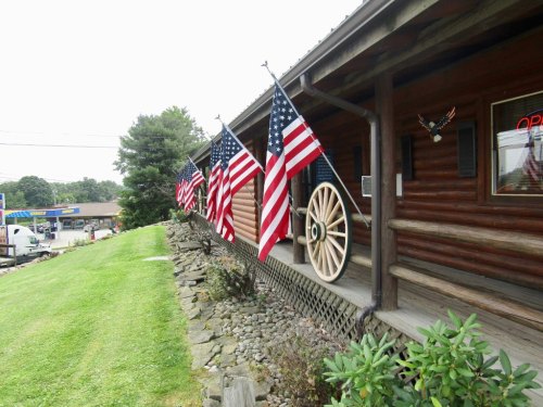 A log cabin with American flags hanging along the porch, surrounded by grass and a stone pathway.