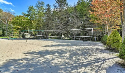 A sandy volleyball court surrounded by trees, with a clear blue sky and colorful autumn foliage in the background.