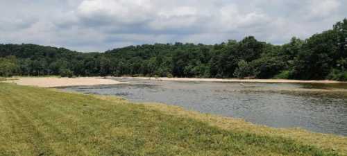 A serene river scene with sandy banks, lush green trees, and a cloudy sky in the background.