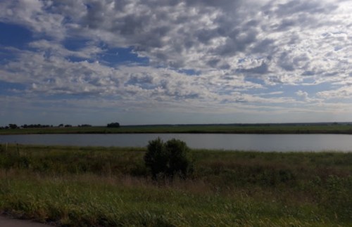 A serene landscape featuring a calm body of water surrounded by lush green fields and a cloudy blue sky.