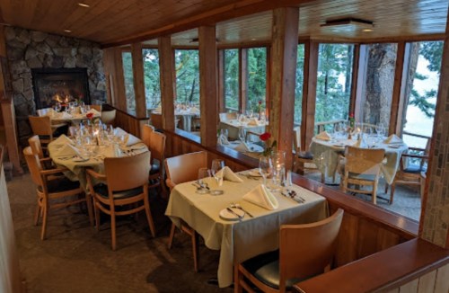 Cozy restaurant interior with elegantly set tables, large windows, and a fireplace, overlooking a scenic view.