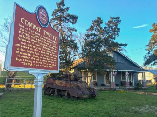 Historical marker for Conway Twitty in front of a house and an old tank, surrounded by trees and grass.