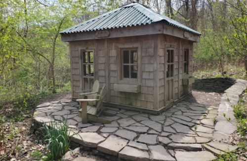 A small wooden cabin with a green metal roof, surrounded by trees and a stone pathway.