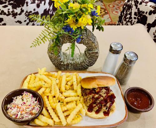 A plate with pulled pork sandwich, crinkle fries, coleslaw, and barbecue sauce, alongside a vase of flowers.