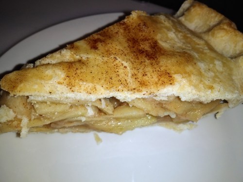 A slice of apple pie on a white plate, featuring a golden crust and cinnamon topping.