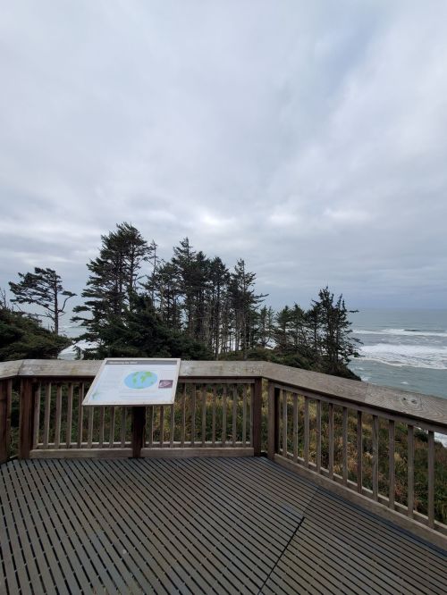 View from a wooden deck overlooking a cloudy ocean scene with trees in the background. Informational sign in foreground.