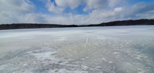 A frozen lake under a cloudy sky, with a snowy landscape and trees in the background.