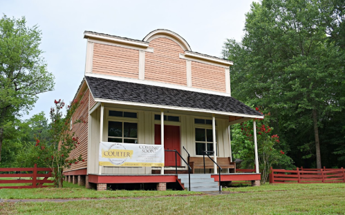 A pink historic building with a black roof, featuring a red door and surrounded by greenery and a wooden fence.
