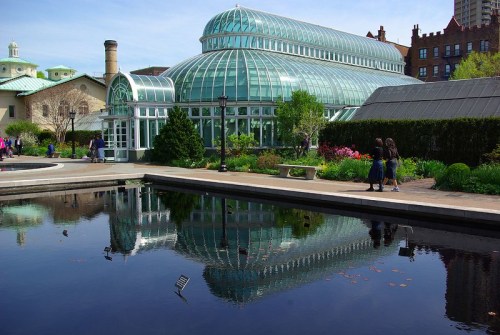 A glass conservatory reflects in a calm pond, surrounded by greenery and people enjoying the garden.