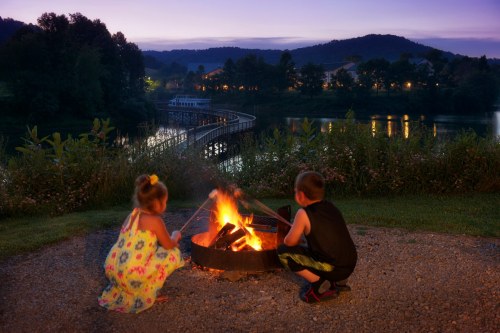 Two children sit by a campfire at dusk, with a lake and mountains in the background.