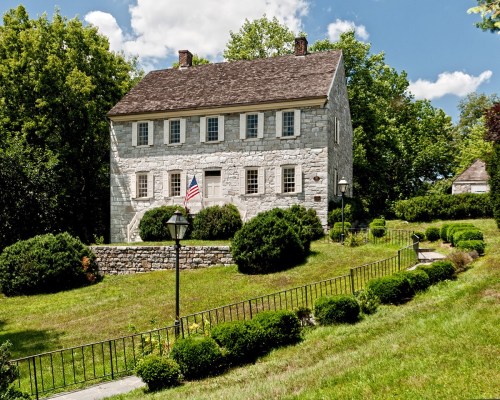 A historic stone house with an American flag, surrounded by lush greenery and a well-maintained garden.