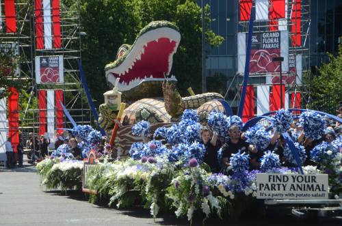 A colorful float shaped like a crocodile, adorned with flowers, in a parade with people holding signs and decorations.