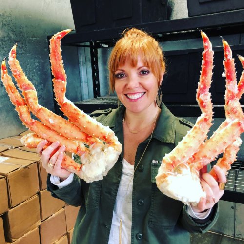 A woman smiles while holding two large crab legs in a storage area filled with boxes.