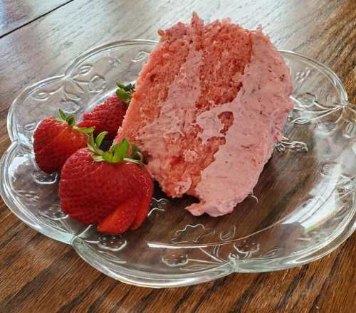 A slice of pink strawberry cake on a glass plate, accompanied by fresh strawberries.