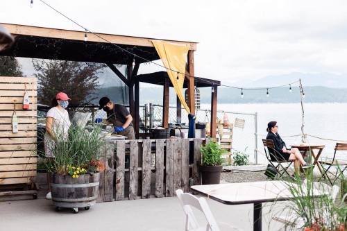 Outdoor café scene by the water, featuring staff in masks, wooden structures, and patrons enjoying the view.