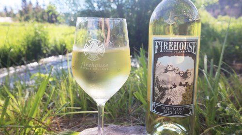 A glass of white wine beside a bottle of Firehouse Rushmore Riesling, set against a lush green background.