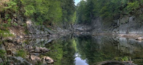 A serene river flows through a rocky canyon, surrounded by lush green trees and reflecting the clear sky above.