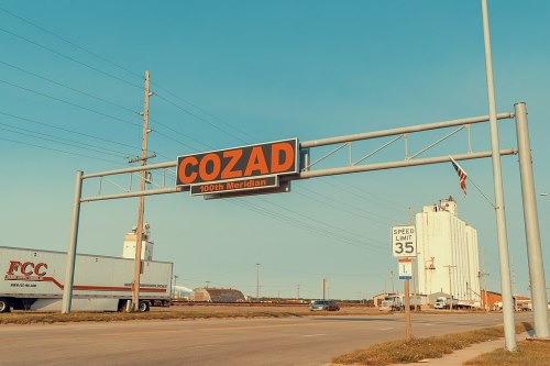 Sign for Cozad, Nebraska, marking the 100th Meridian, with trucks and grain elevators in the background.