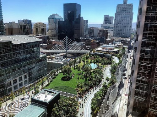 Aerial view of a vibrant urban park surrounded by skyscrapers and city buildings on a sunny day.