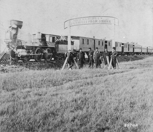 Historic black-and-white photo of a steam locomotive and train cars, with men posing near a distance sign.