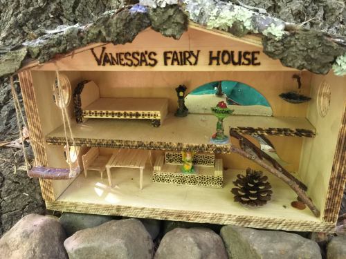 A miniature fairy house with wooden furniture, a blue window, and a sign reading "Vanessa's Fairy House."