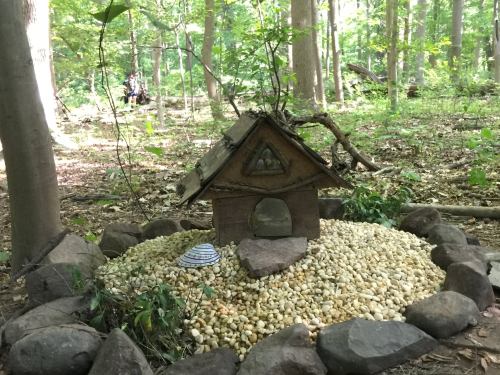 A small wooden house surrounded by pebbles and stones, set in a forested area with trees in the background.