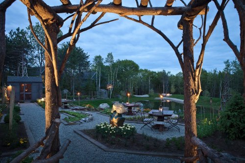A serene garden at dusk, featuring a wooden arch, stone paths, tables, and a pond surrounded by trees.