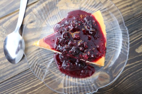 A slice of cheesecake topped with a rich berry sauce on a clear glass plate, with a spoon beside it.