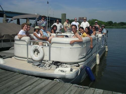 A group of people enjoying a sunny day on a boat, docked by a wooden pier near a river.