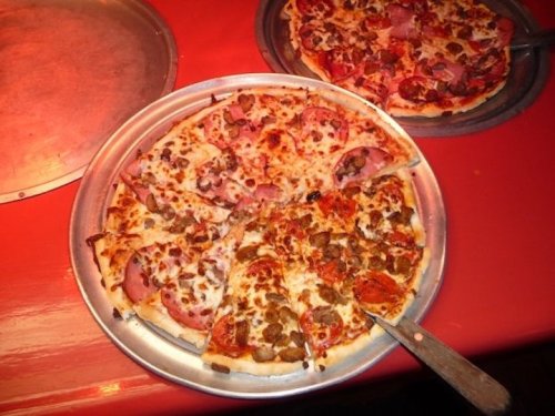 Two pizzas on metal trays: one topped with ham and cheese, the other with pepperoni and sausage, on a red table.
