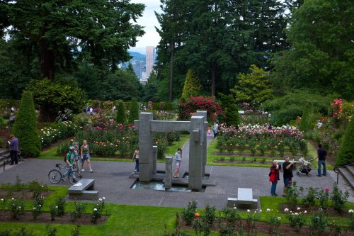 A landscaped garden with a modern sculpture, surrounded by blooming roses and visitors enjoying the scenery.