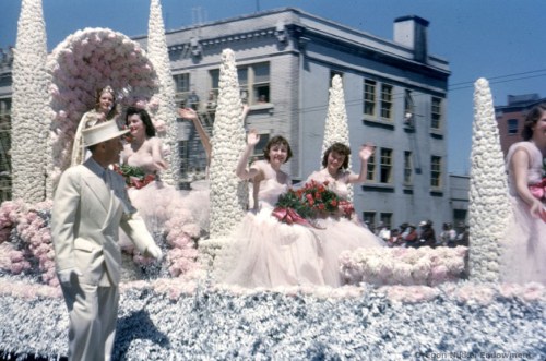 A colorful parade float adorned with flowers, featuring smiling women in dresses, waving to the crowd.