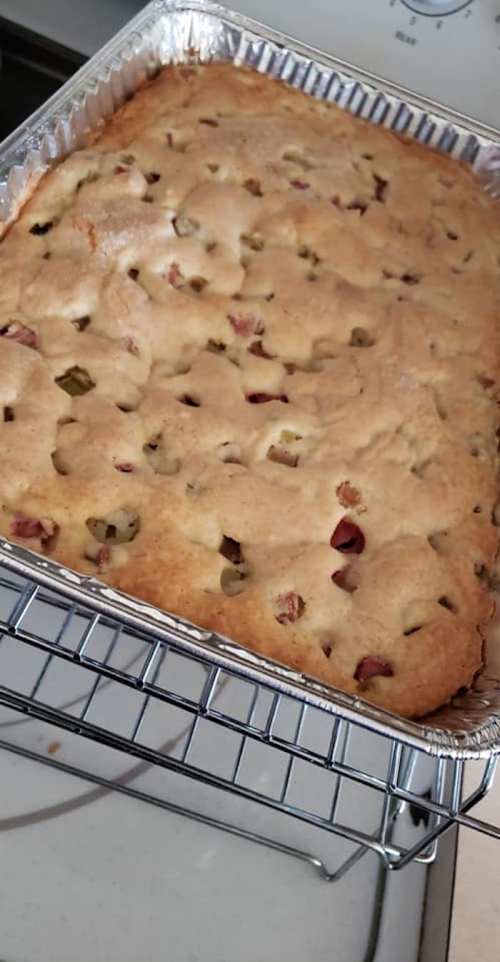 A freshly baked cake in a silver foil pan, cooling on a wire rack, with a golden-brown top and visible fruit pieces.