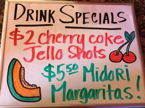 A whiteboard sign listing drink specials: $2 cherry coke jello shots and $5.50 Midori margaritas, with colorful drawings.